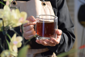 A person in a beige apron holding a clear glass mug filled with a dark amber-brown liquid, with soft-focus yellow flowers on the left and a bright, warm background.