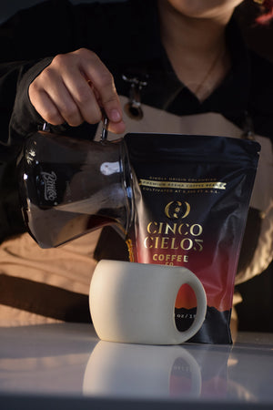 A person pours coffee from a glass carafe into a ceramic mug next to a bag of Cinco Cielos coffee, set against a dark background.