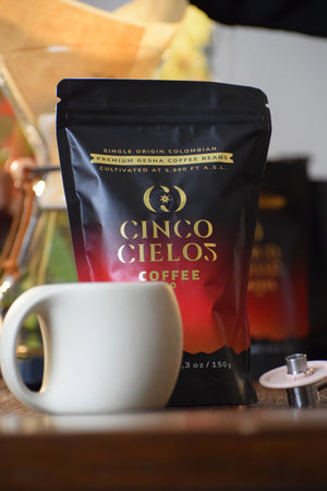 A bag of Cinco Cielos Coffee beans in front of a white ceramic mug on a wooden surface, with a coffee maker blurred in the background.