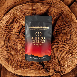 A stand-up pouch of Cinco Cielos Coffee Co. single-origin Colombian coffee, featuring a black-to-red gradient design with gold lettering, placed on a rustic wooden log slice background.